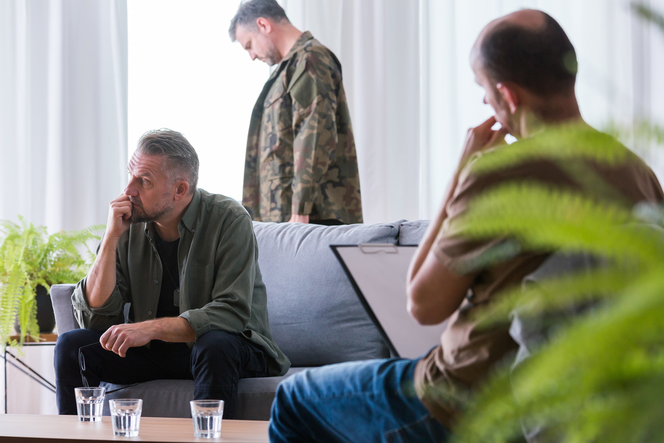 Tres hombres participan en una sesión de terapia grupal en un entorno tranquilo, con uno de ellos vestido con un uniforme militar, simbolizando el tratamiento del trauma y el estrés postraumático relacionado con experiencias difíciles como las vividas en el servicio militar.