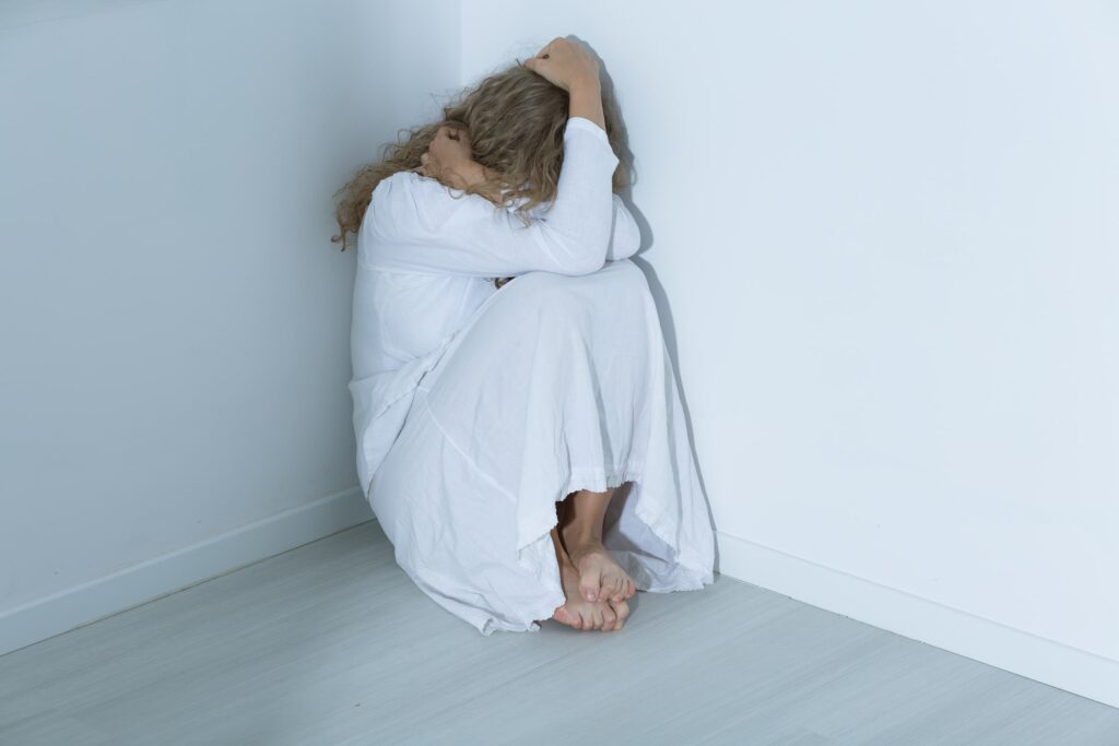 Mujer sentada en el suelo, encogida en una esquina con las manos sobre la cabeza, simbolizando el impacto emocional de las causas y factores de riesgo relacionados con los trastornos de ansiedad.