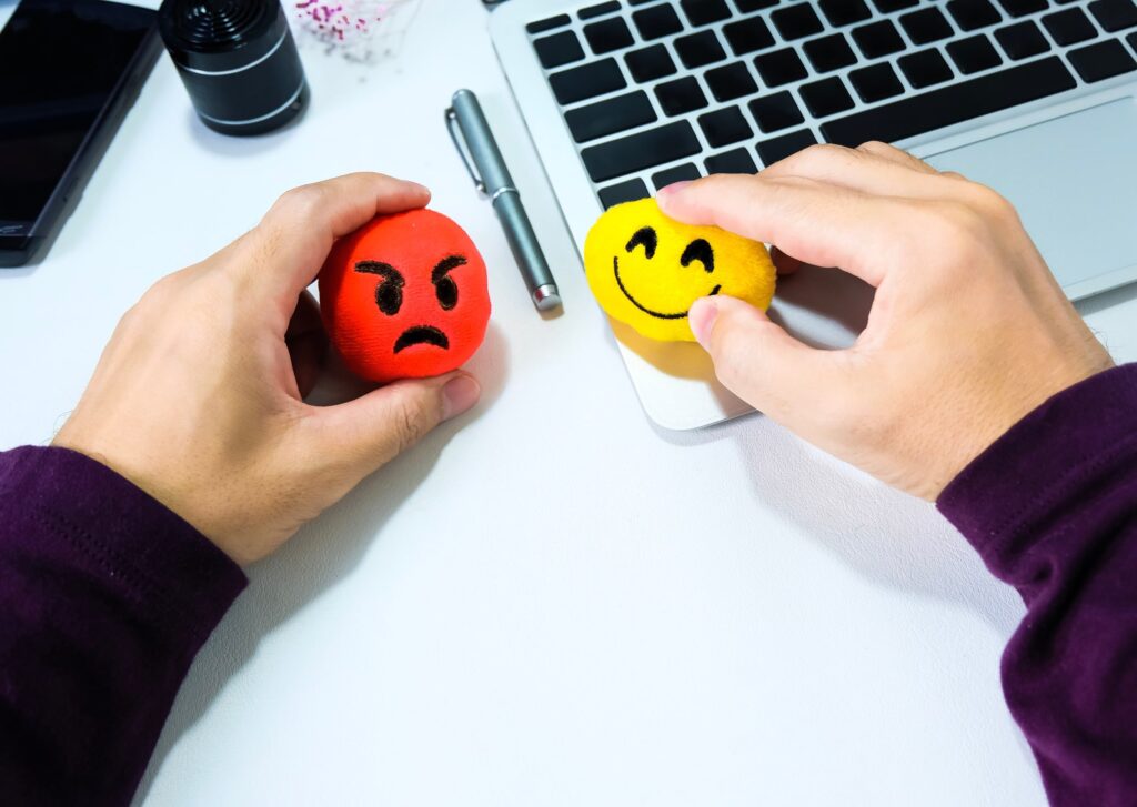 Dos manos sostienen pelotas antiestrés: una roja con expresión de enfado y otra amarilla con expresión de felicidad, sobre un escritorio con un ordenador portátil. Representan estrategias para gestionar emociones y resolver conflictos en terapia de pareja.